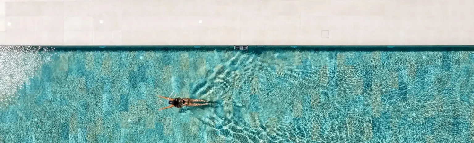 Vista a&eacute;rea de una persona nadando junto al borde de la piscina, mostrando materiales para bordes de piscinas en tono claro